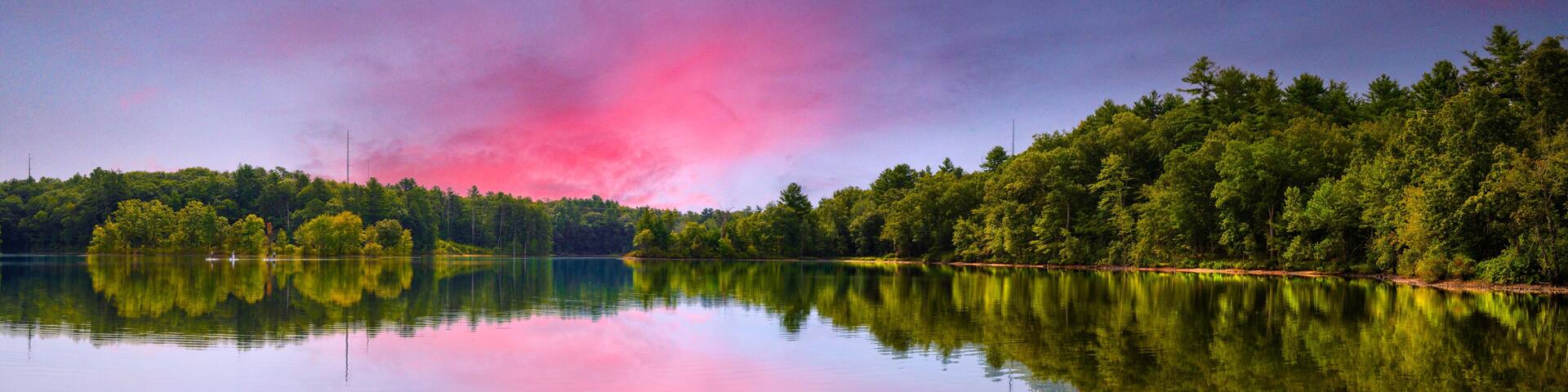 A calm lake with dense forest on one side and a clear sky with pink clouds.