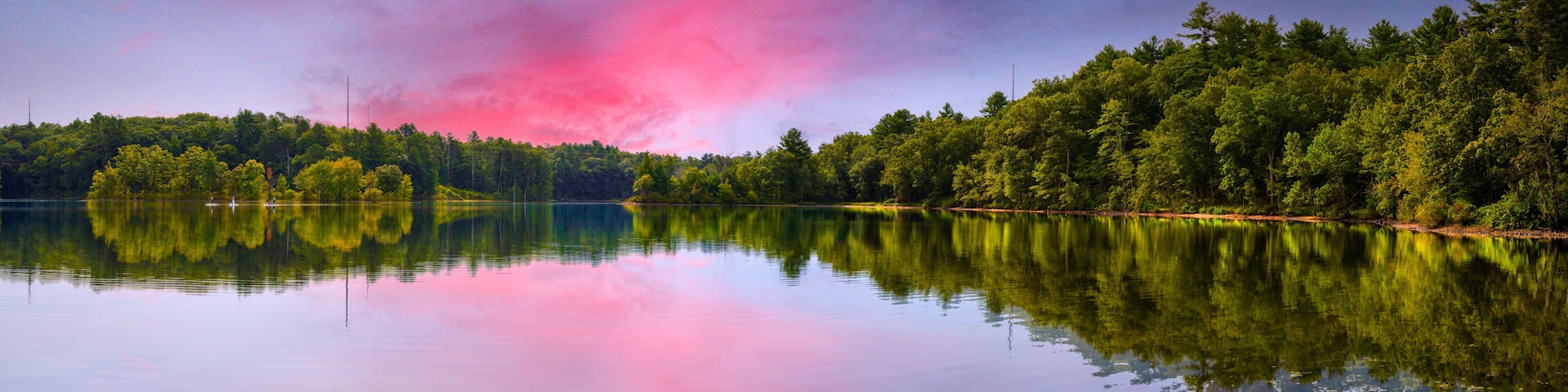 Un lago tranquilo con un bosque denso a un lado y un cielo despejado con nubes rosadas.