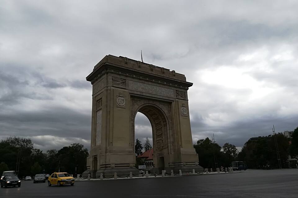 A large stone arch monument with inscriptions and a clock, surrounded by trees and vehicles on a cloudy day.
