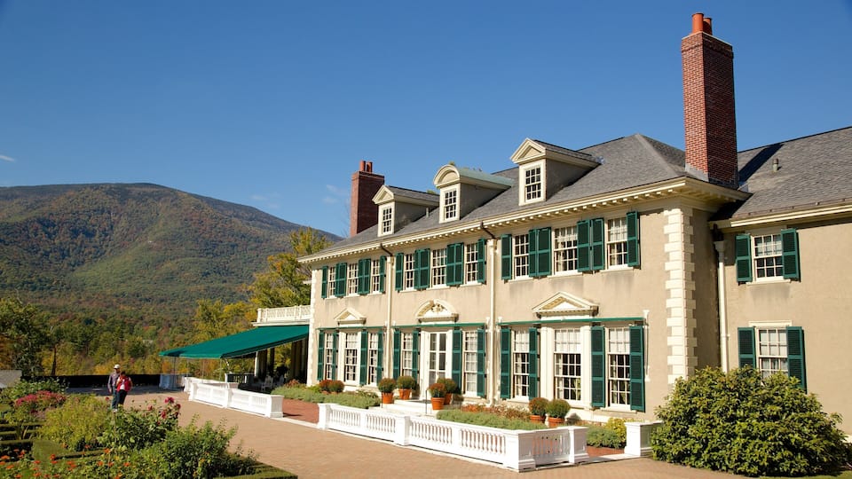 A large, historic building with green shutters and a brick chimney, surrounded by lush gardens and a mountainous backdrop.