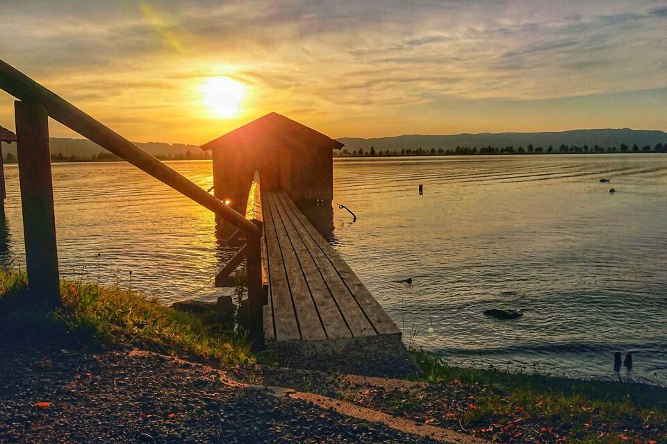Un muelle de madera se extiende sobre un lago tranquilo al atardecer, con una cadena montañosa en la distancia.