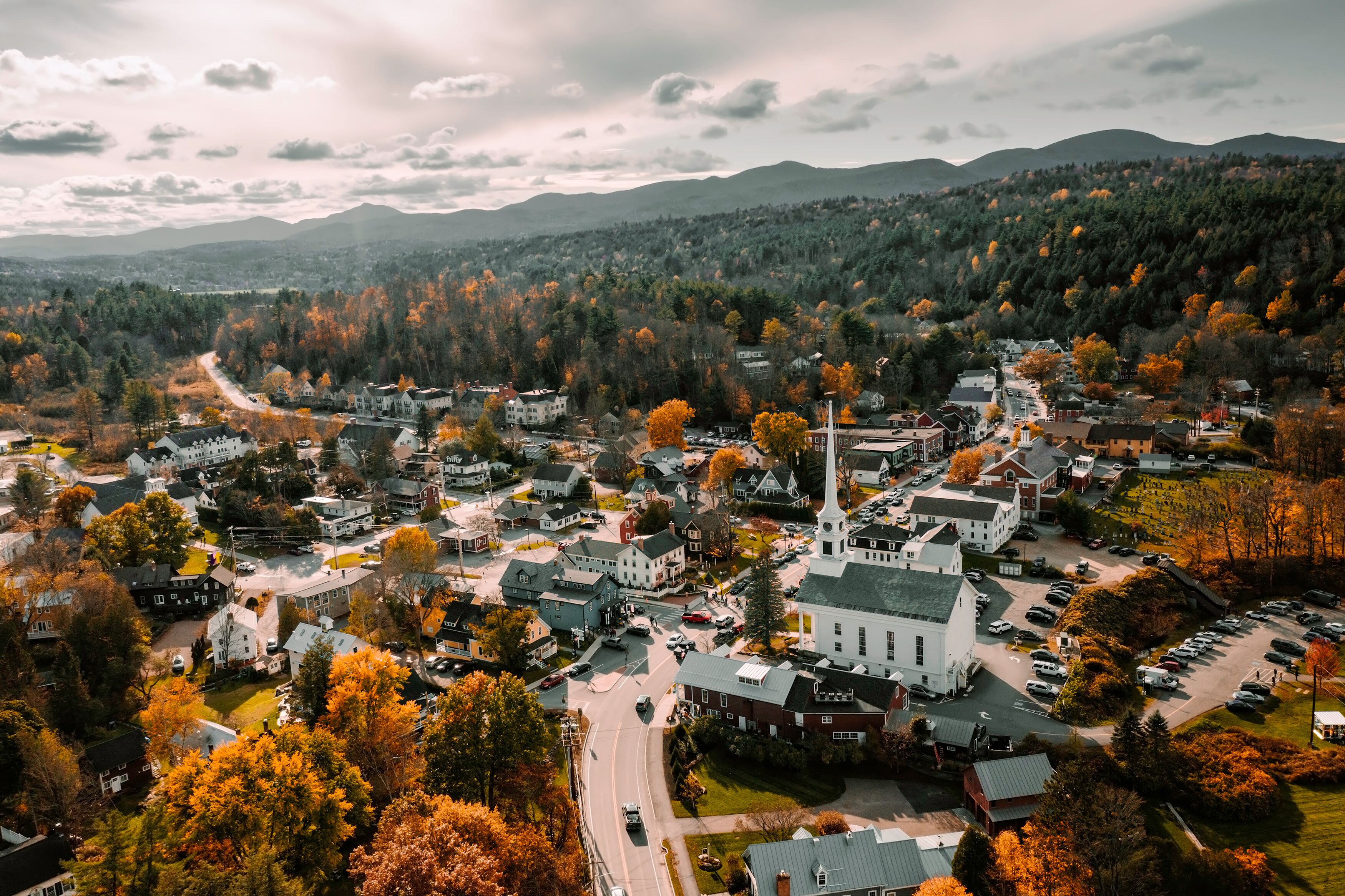 A town with a prominent church, surrounded by autumn-colored trees and a mountain range in the background.