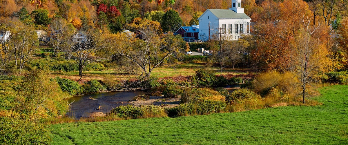 Une église blanche, dotée d’un clocher, entourée d’un feuillage automnal et bordée par une rivière.