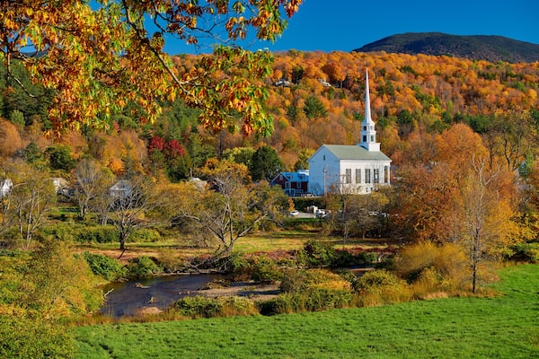 Une église blanche, dotée d’un clocher, entourée d’un feuillage automnal et bordée par une rivière.