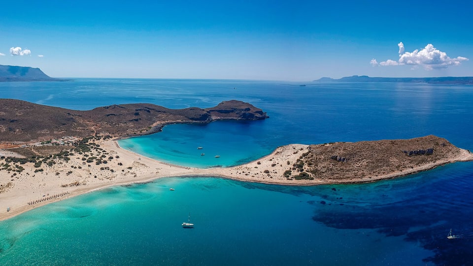 A coastal landscape with a beach, clear turquoise waters, and distant mountains.