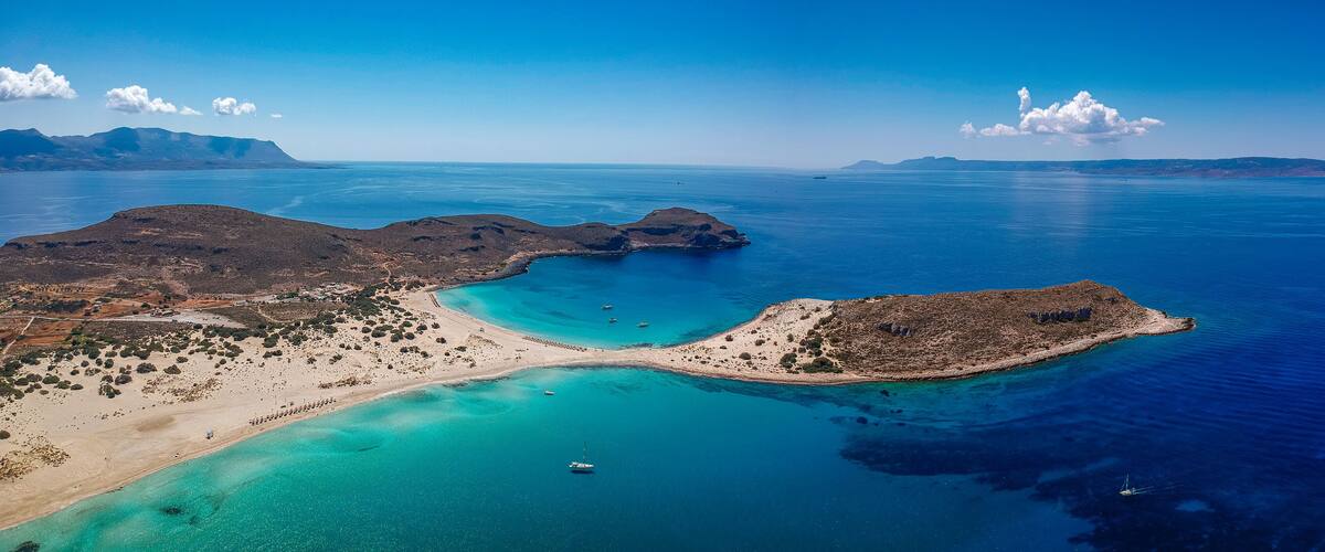 A coastal landscape with a beach, clear turquoise waters, and distant mountains.