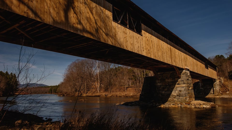 A wooden bridge over a river with stone supports and a forested landscape in the background.