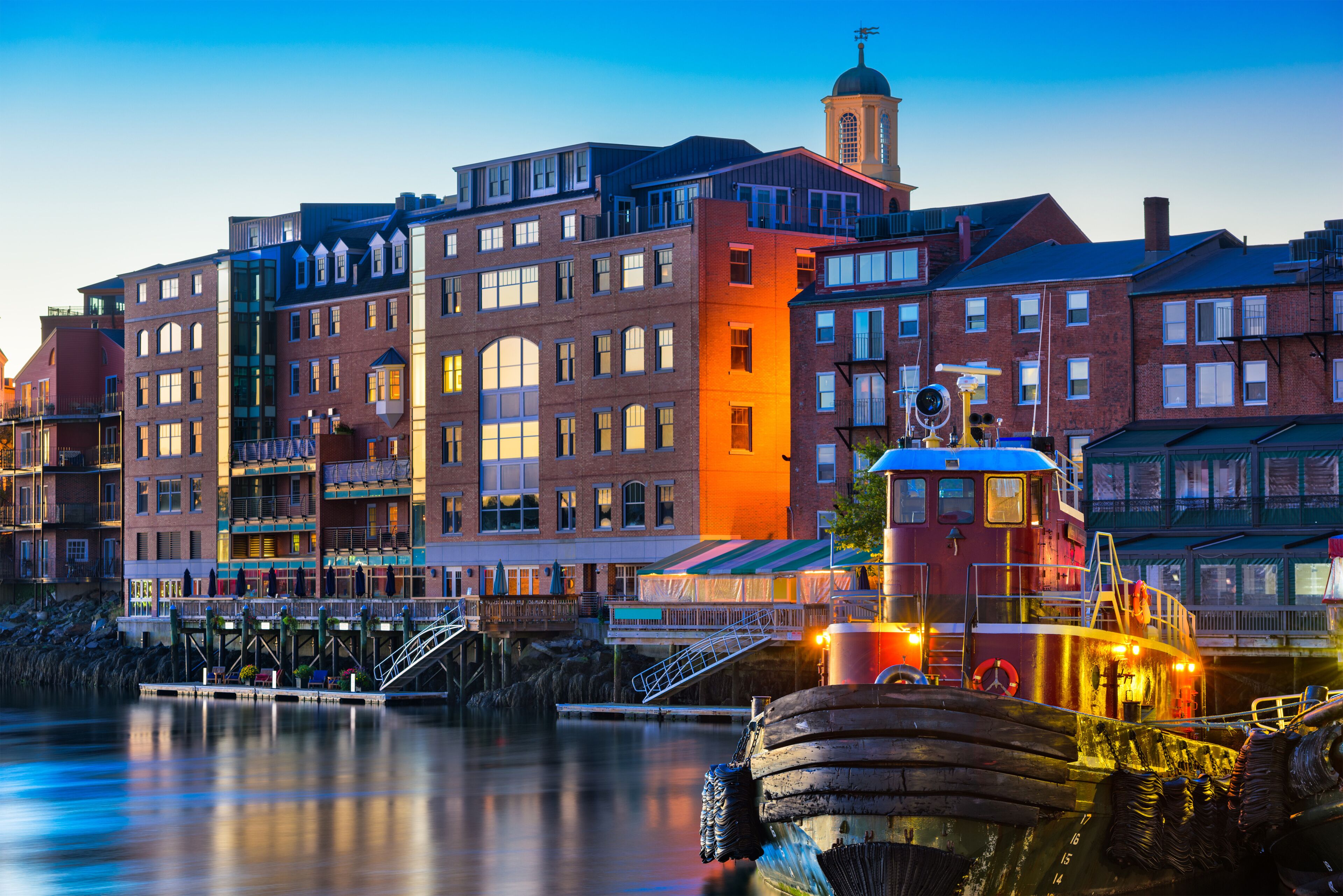 A waterfront cityscape with a historic building featuring a clock tower, a modern red building, and a traditional boat docked at the pier.