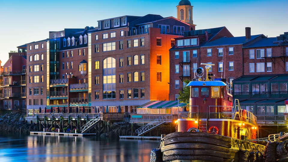 A waterfront cityscape with a historic building featuring a clock tower, a modern red building, and a traditional boat docked at the pier.