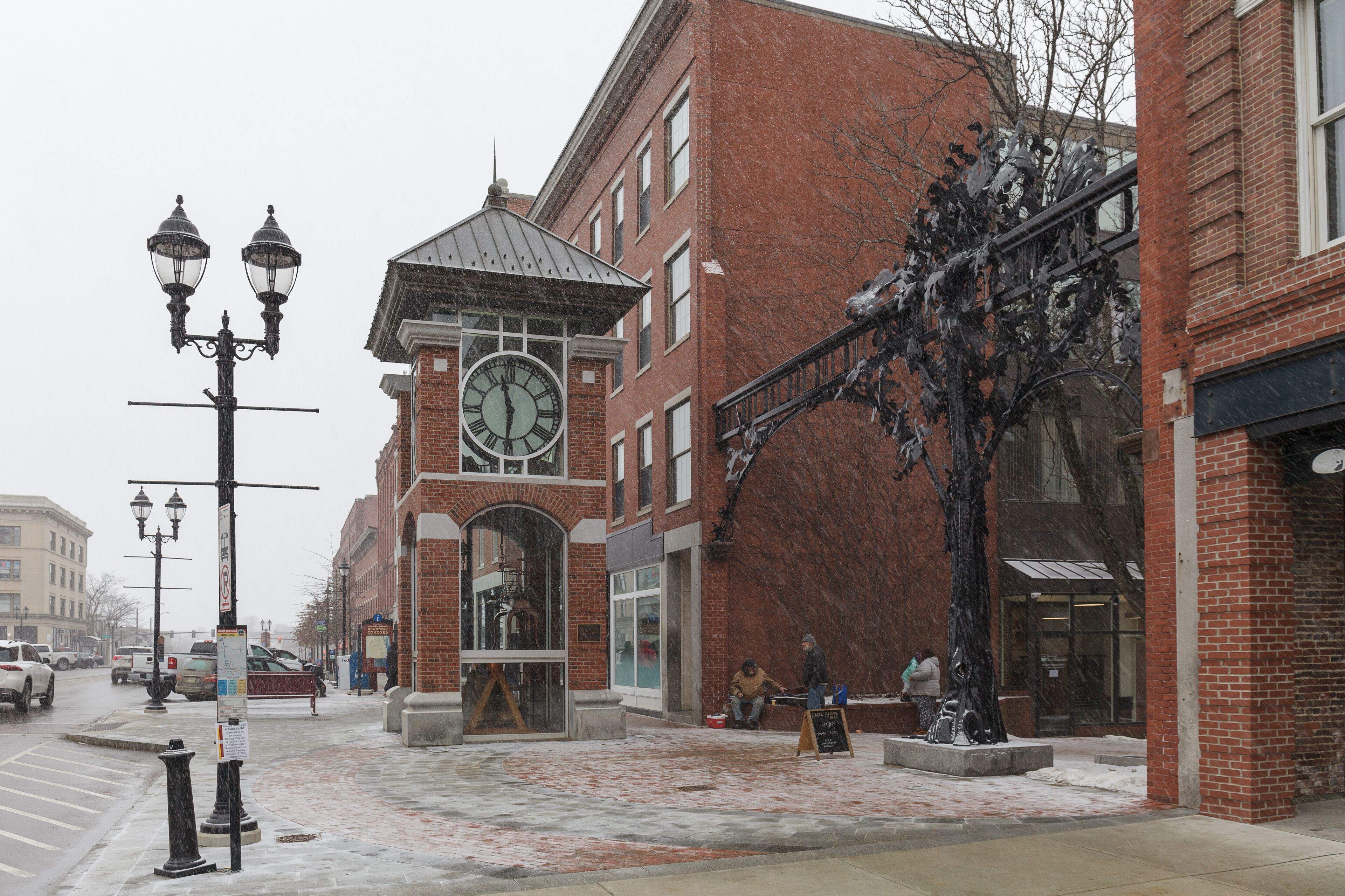 A brick building with a clock tower, a street with cars, and a snow-covered sidewalk.