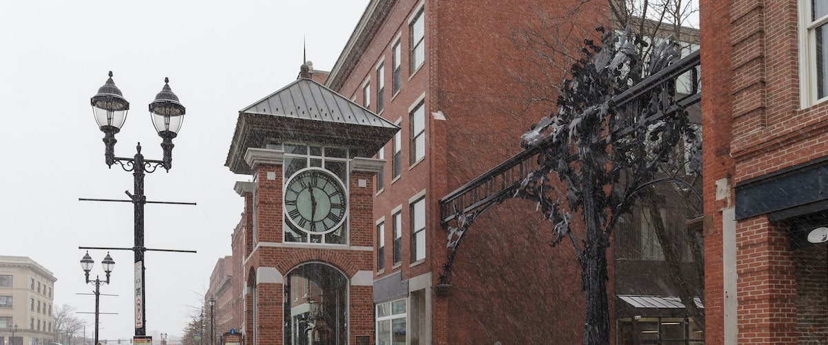 A brick building with a clock tower, a street with cars, and a snow-covered sidewalk.