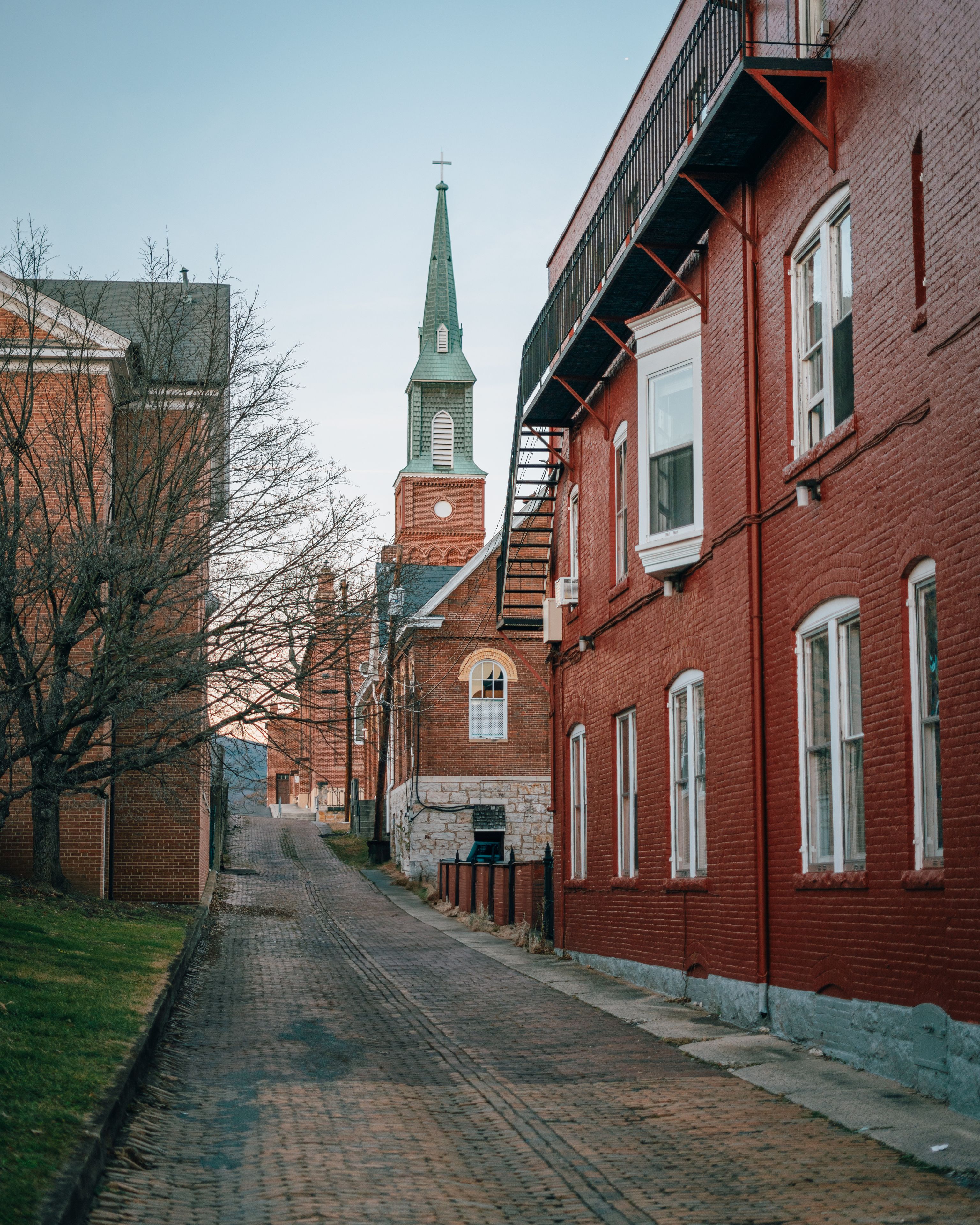 A cobblestone street lined with red brick buildings, a church with a green spire, and bare trees.