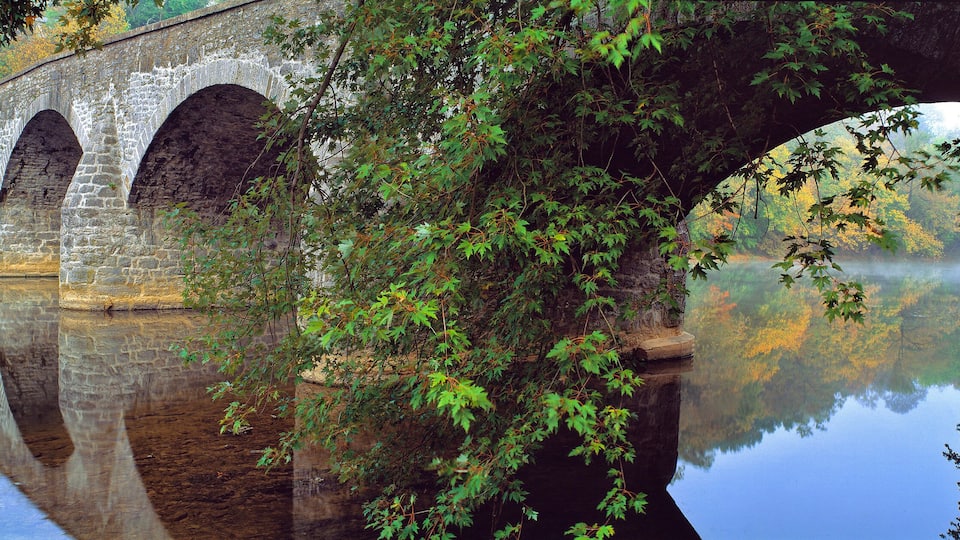 A stone bridge with multiple arches over a calm river, surrounded by lush greenery and trees reflecting in the water.