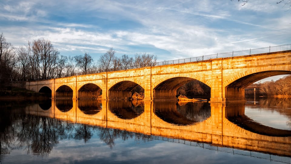 A stone bridge with multiple arches over a calm river, surrounded by bare trees and a clear sky.