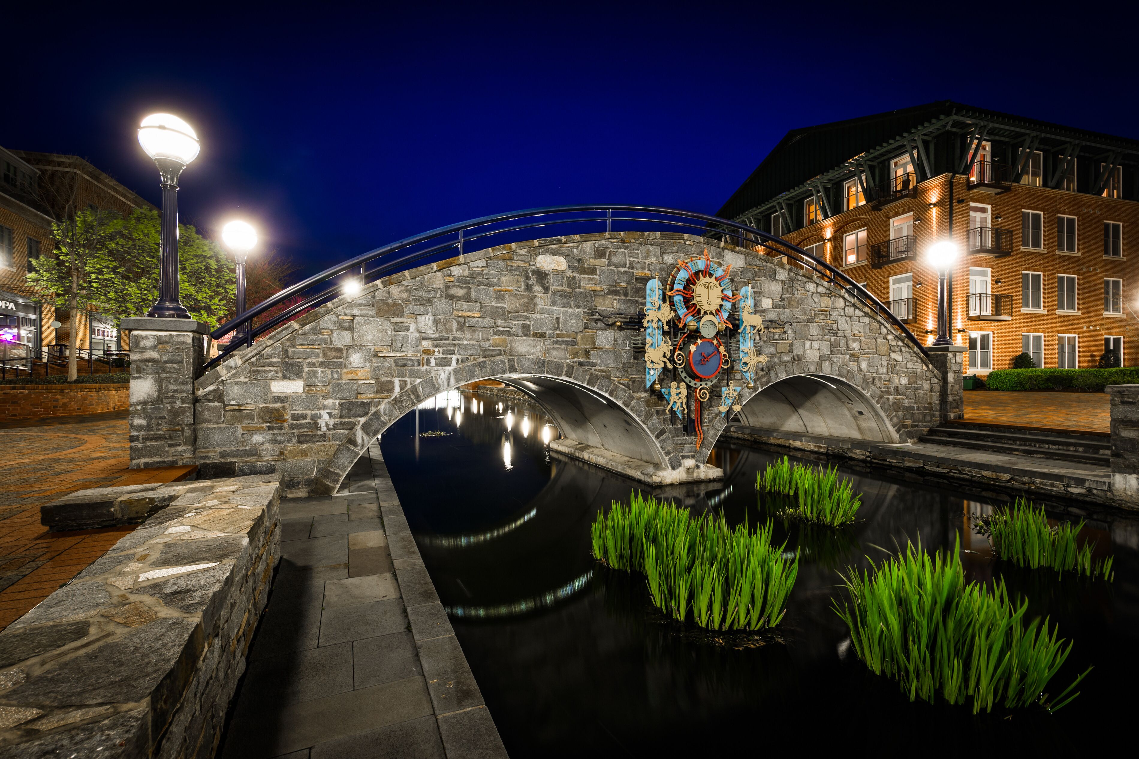 A stone bridge over a canal with a decorative clock, surrounded by buildings and street lamps.