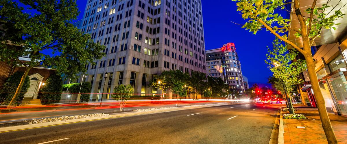 A cityscape at dusk with a prominent high-rise building, a tree-lined street, and a busy road with light trails.