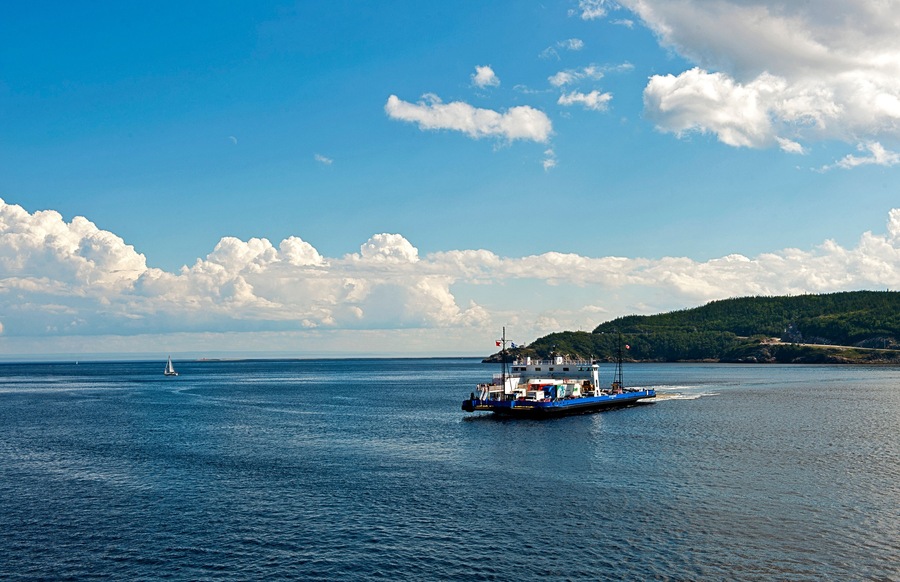 A blue ferry on a calm sea with a sailboat in the distance and a forested island in the background.