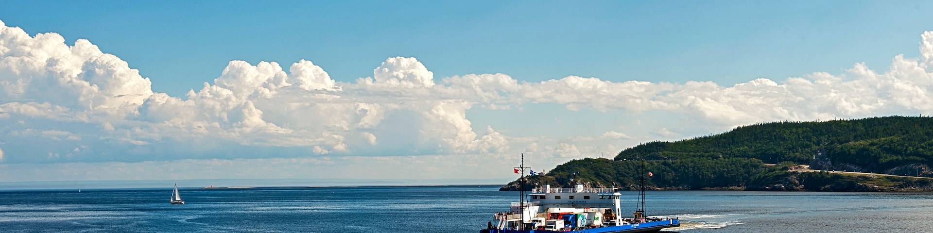 A blue ferry on a calm sea with a sailboat in the distance and a forested island in the background.