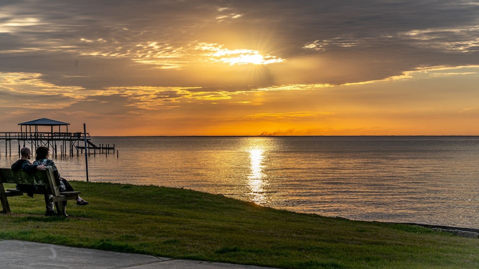 A sunset over a calm body of water with a pier and a bench where two people are sitting.