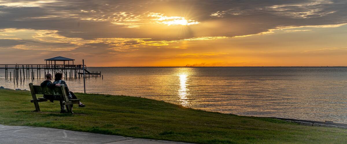 A sunset over a calm body of water with a pier and a bench where two people are sitting.