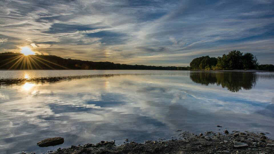 A calm lake with the sun reflecting on the water, surrounded by trees and a clear sky.