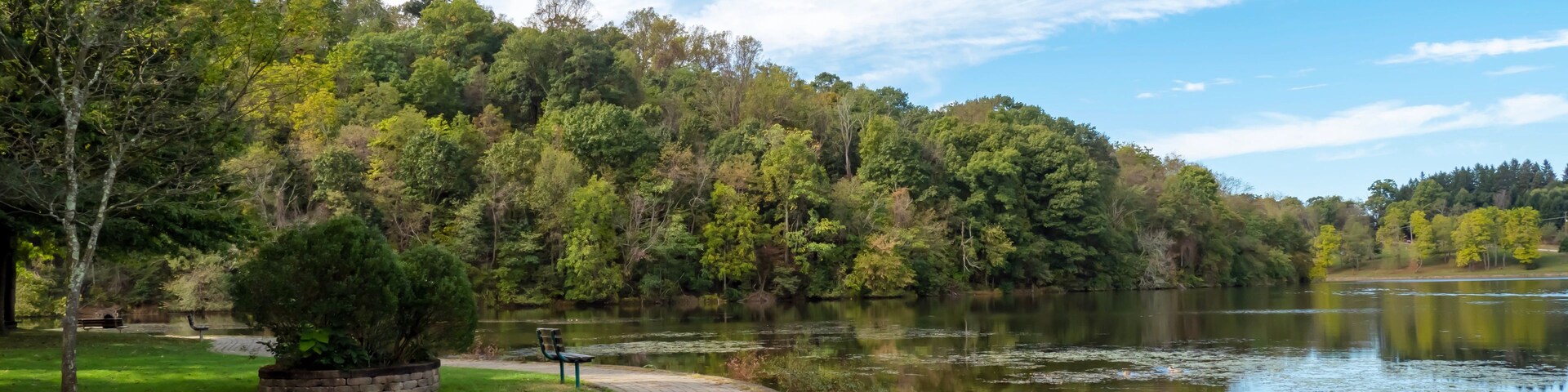 Uma passarela pavimentada ao lado de um lago tranquilo, rodeada por árvores verdes e um céu azul límpido.