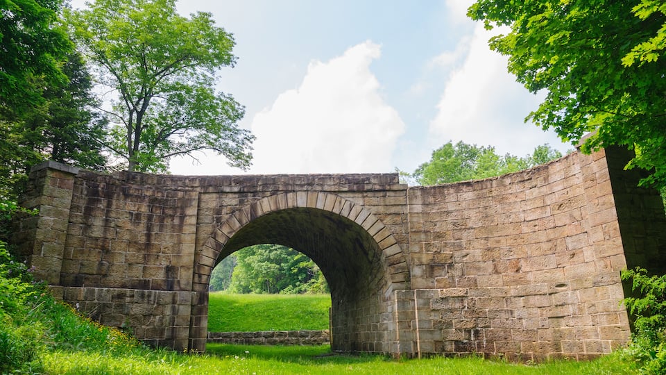 An old stone bridge arching over a grassy field with trees in the background.