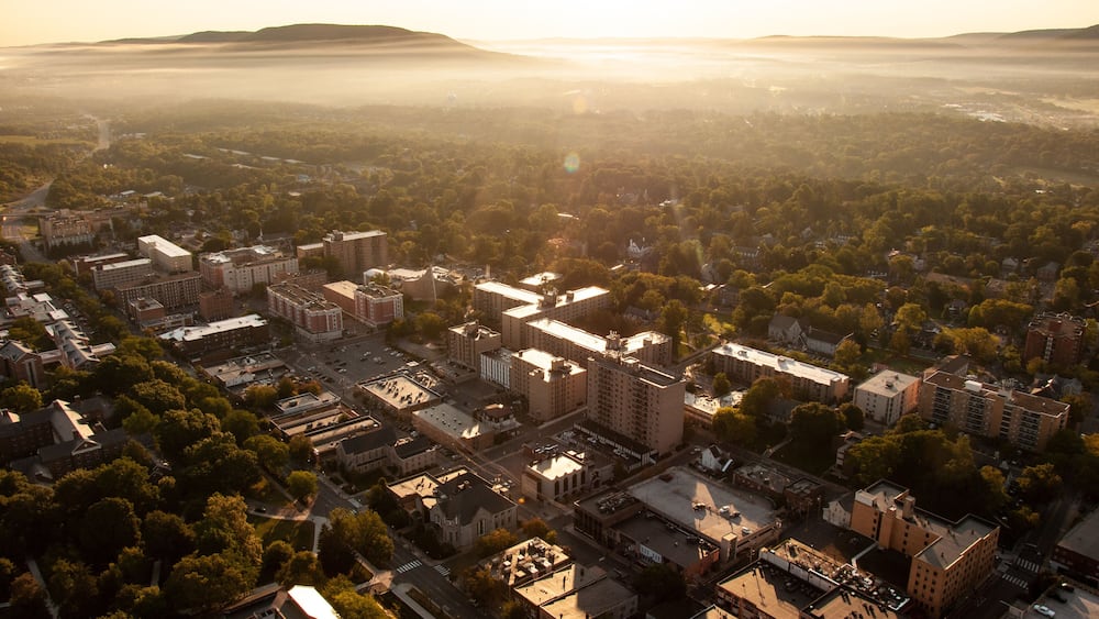 An aerial view of a city with dense urban development, surrounded by green areas and hills in the distance.