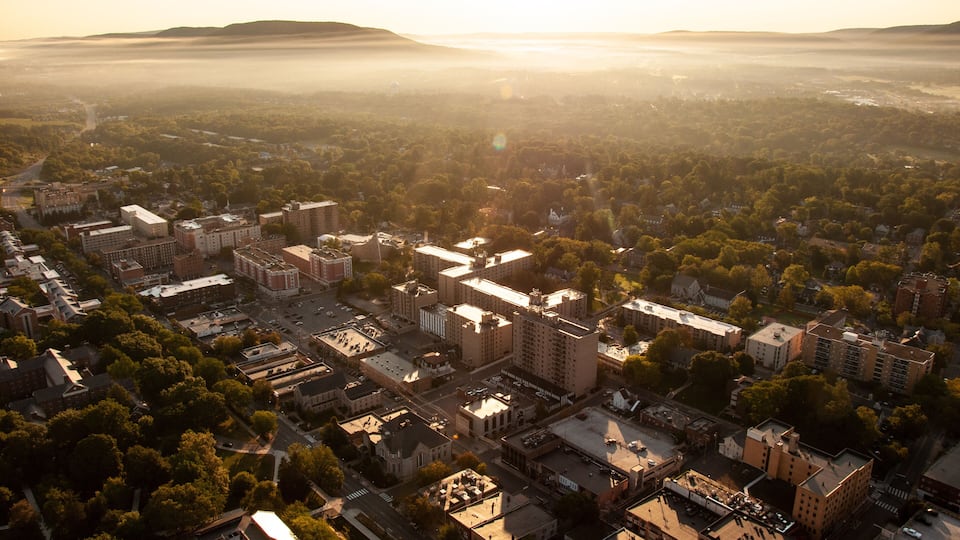 An aerial view of a city with dense urban development, surrounded by green areas and hills in the distance.