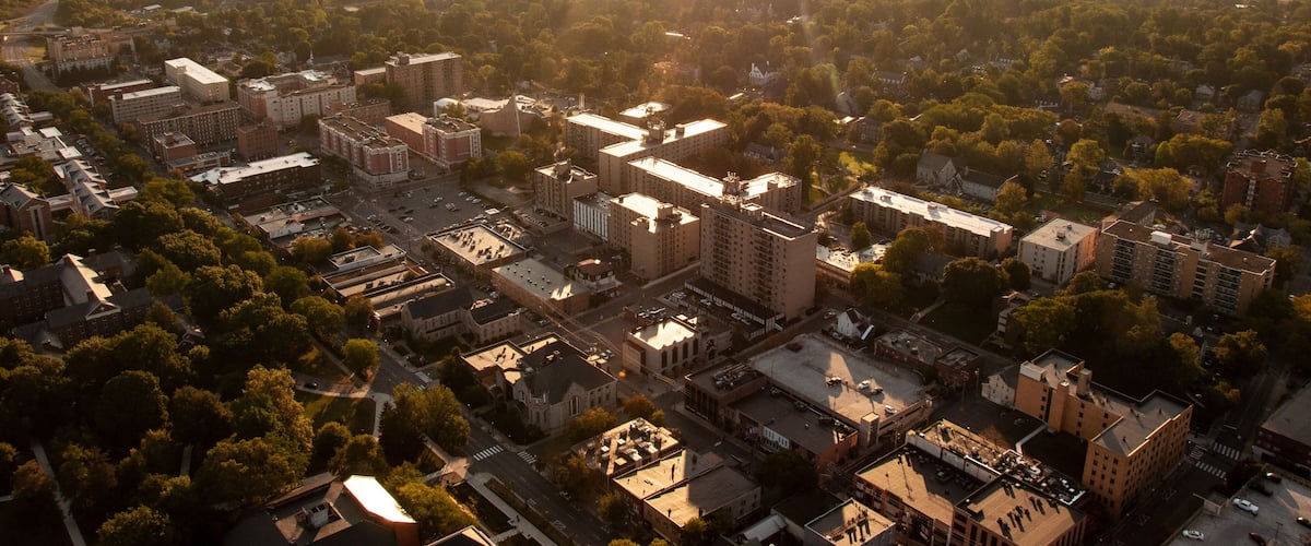 An aerial view of a city with dense urban development, surrounded by green areas and hills in the distance.