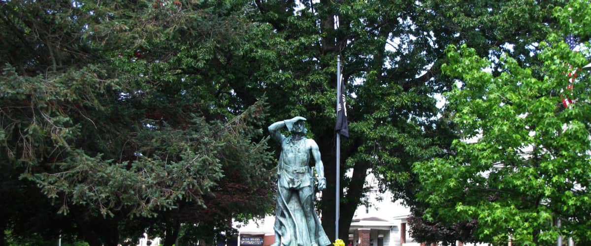 A statue of Christopher Columbus in a park with trees and a building in the background.