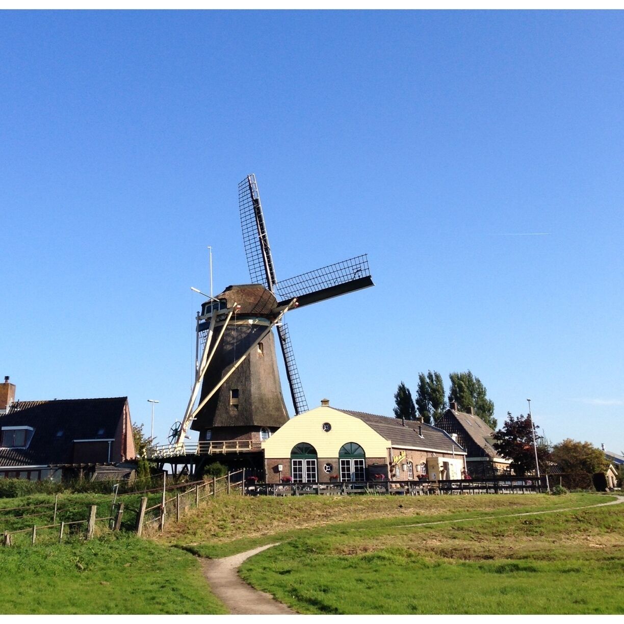 A traditional windmill with a large sail, a yellow building with arched windows, and a grassy field with a path.