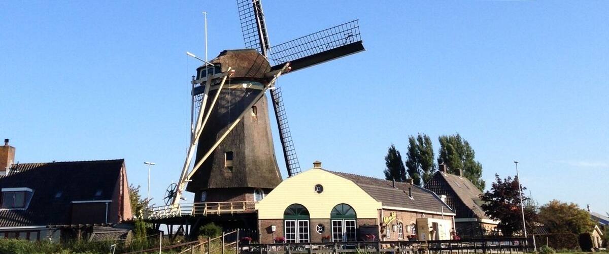 A traditional windmill with a large sail, a yellow building with arched windows, and a grassy field with a path.