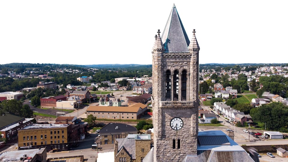 A clock tower with a pointed roof and a clock face, surrounded by a town with various buildings and a river in the background.