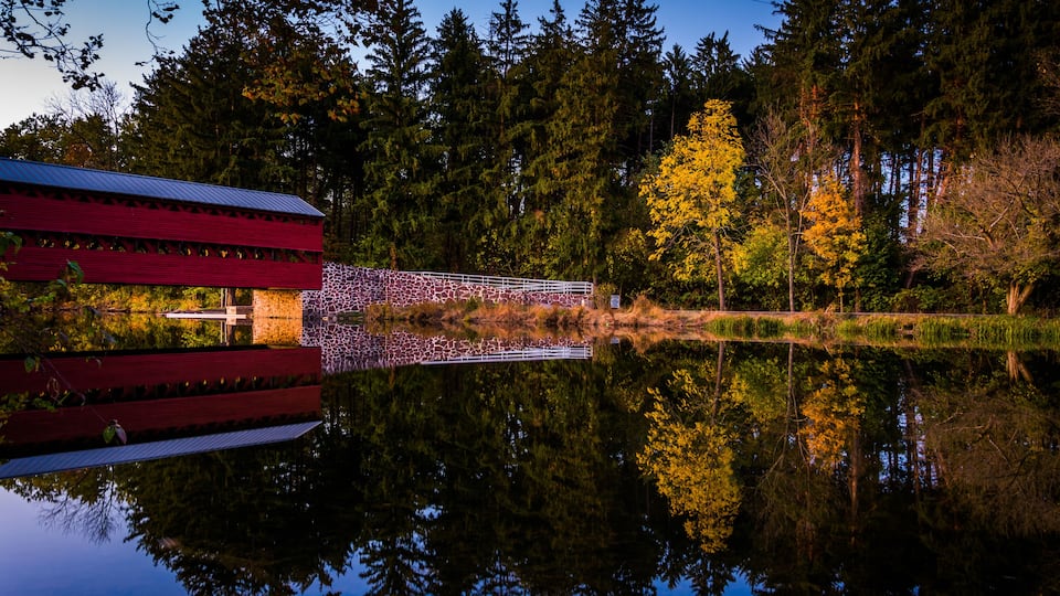 A red covered bridge over a calm lake with reflections of trees and a clear sky.