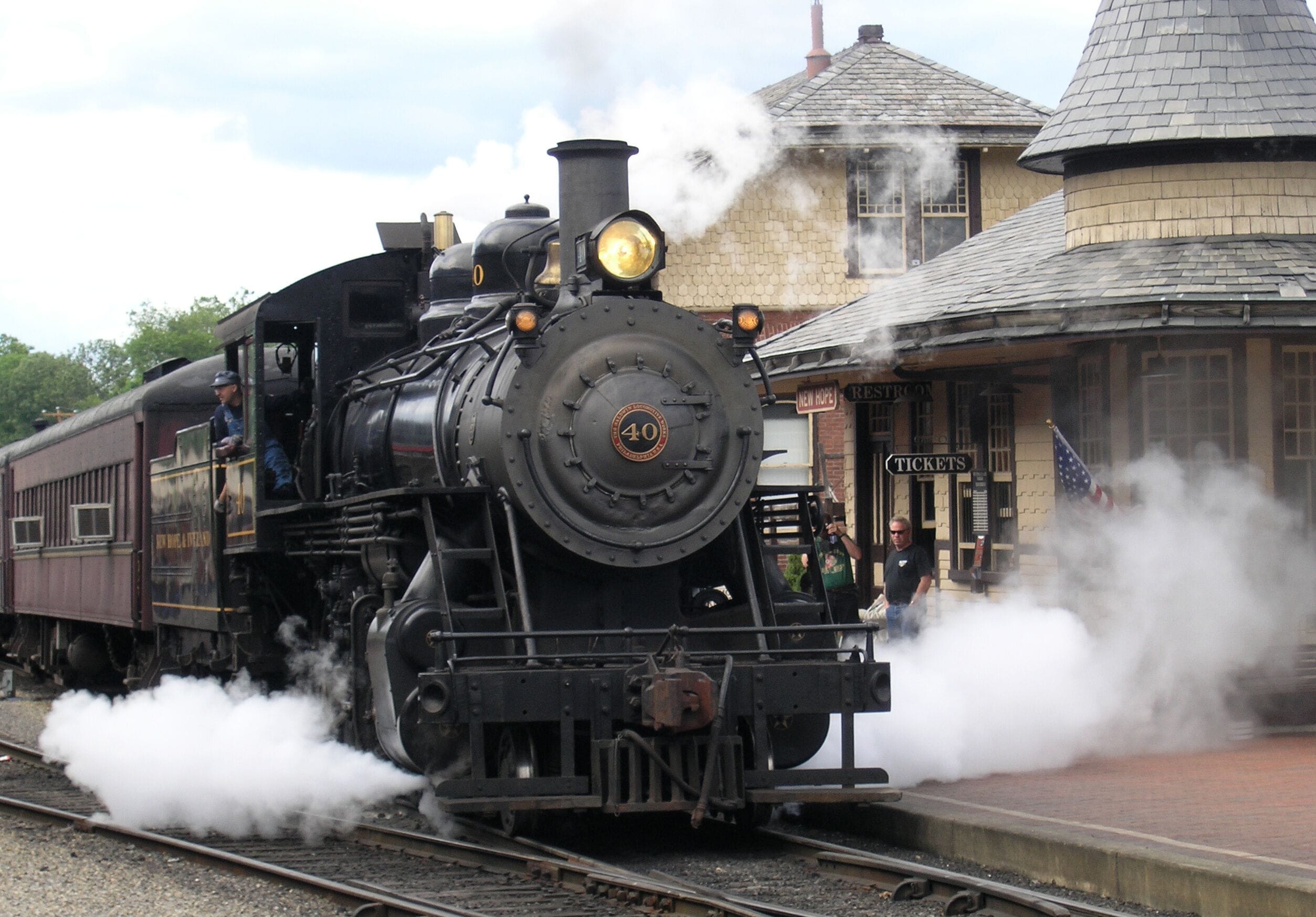 A steam locomotive with the number 40 on its front, emitting steam, at a train station with a brick building and a clock tower in the background.