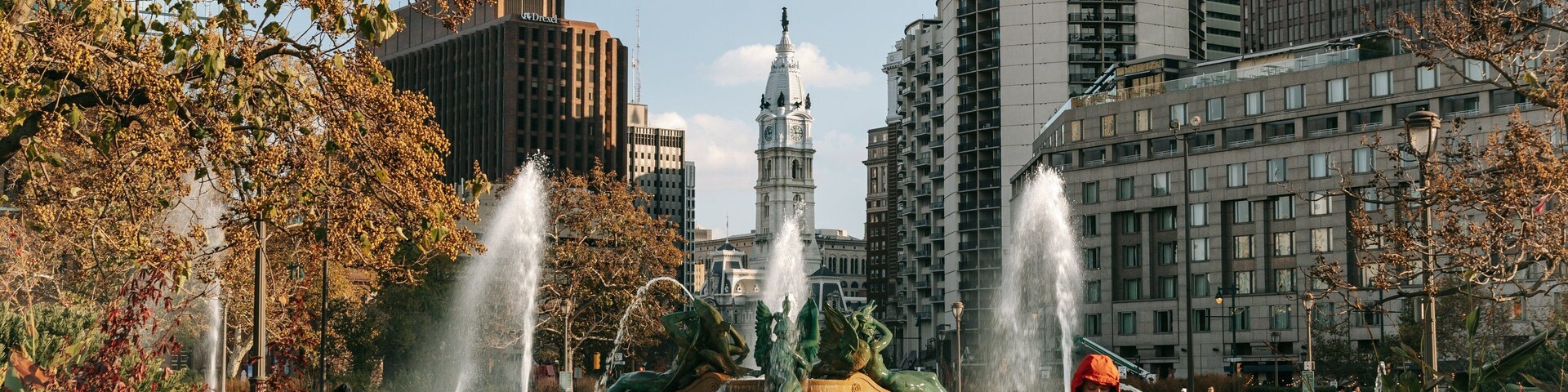 Um parque com fontes, uma torre do relógio e edifícios modernos.