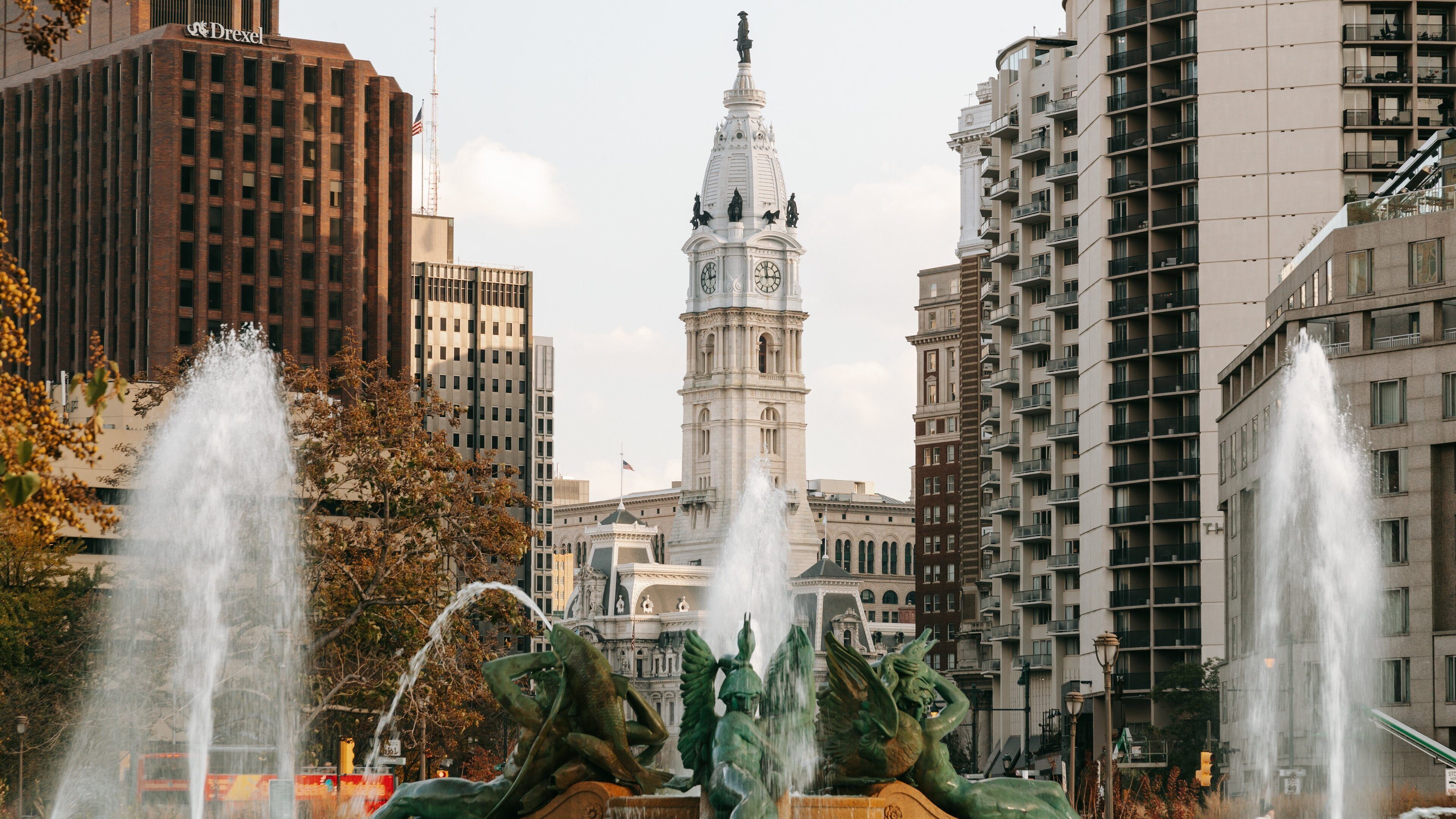 A cityscape with a prominent clock tower, modern high-rise buildings, and a fountain with sculptures.