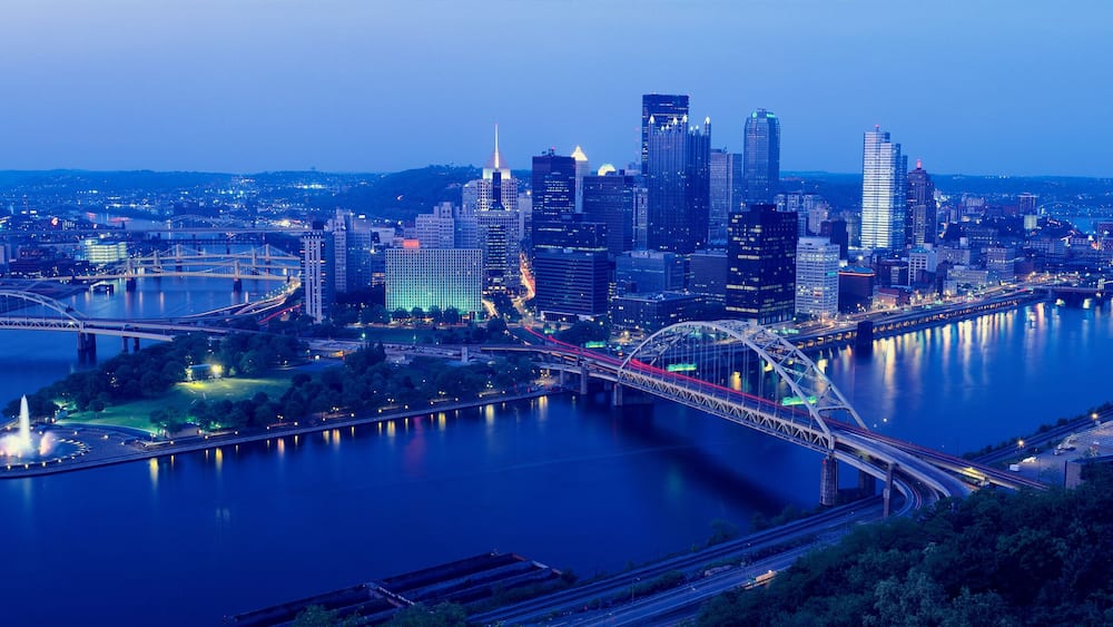 A city skyline with a river, bridges, and a park with a fountain.