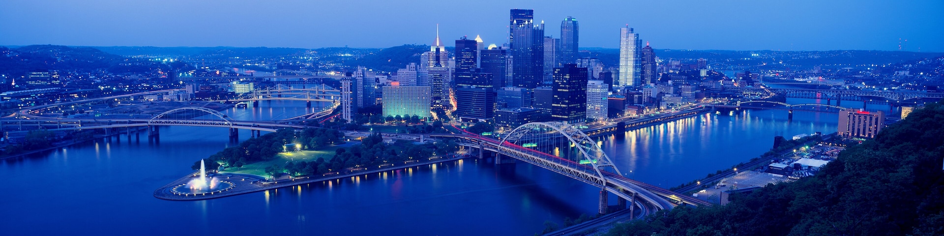 A city skyline with a river, bridges, and a park with a fountain.