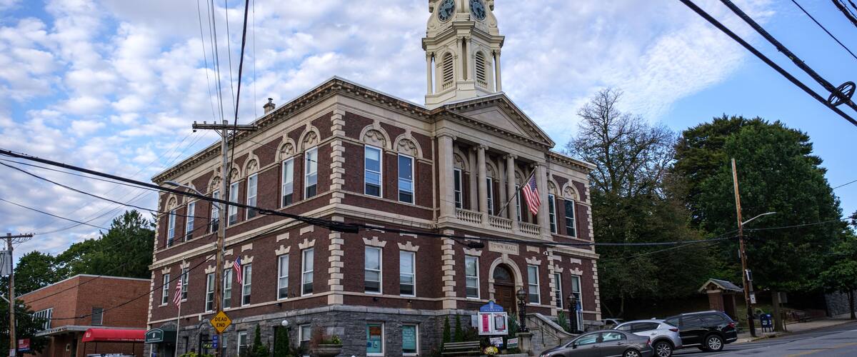 A historic building with a clock tower, a town hall sign, and a street with cars and a bench.