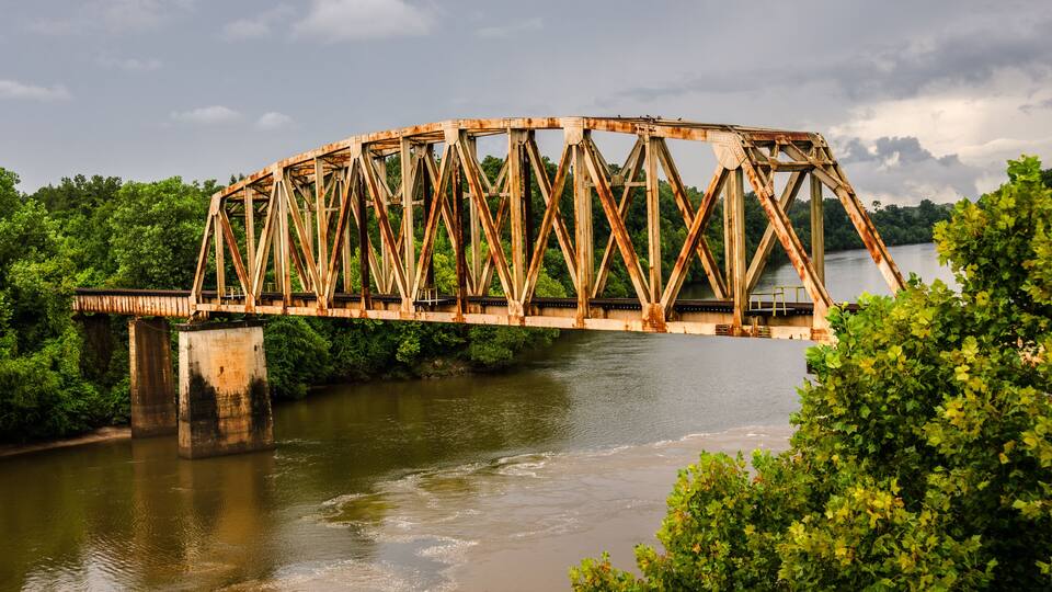A rusted metal bridge over a river with lush greenery on both sides.