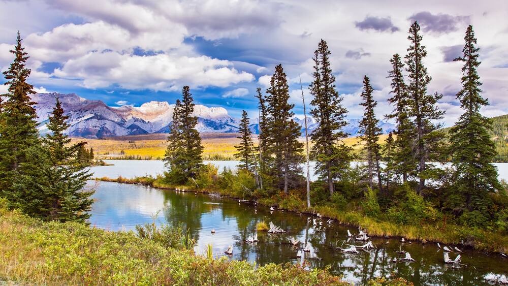 A mountain landscape with a lake, trees, and a cloudy sky.