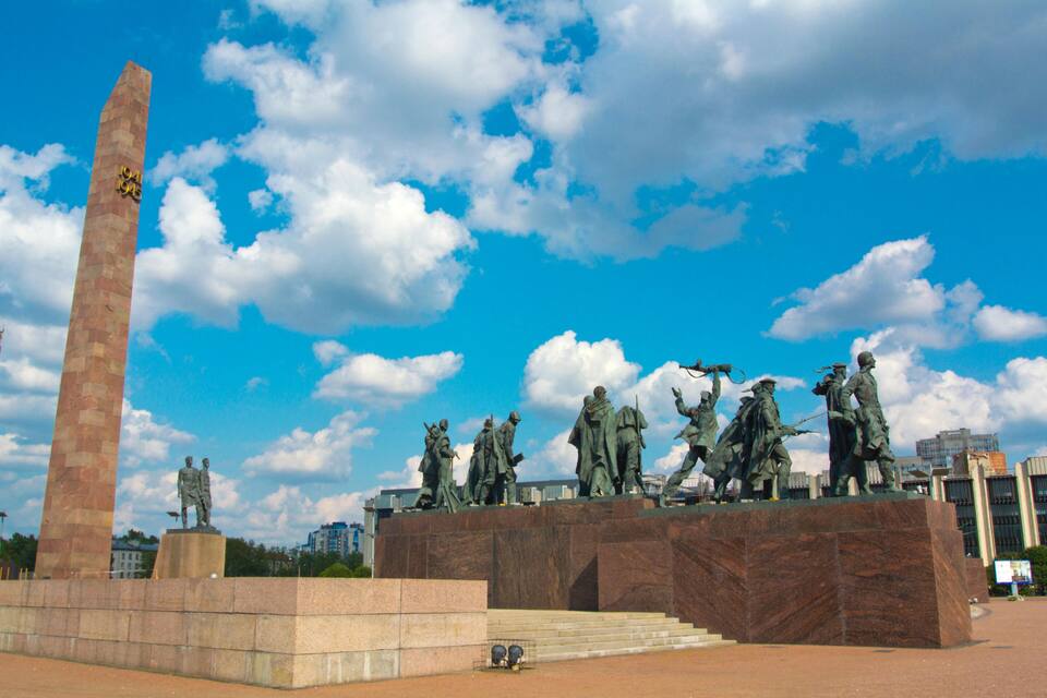 A monument with statues and a tall obelisk under a blue sky with clouds.