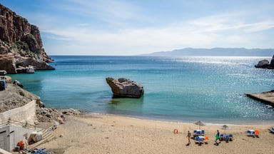 A beach with clear turquoise water, a large rock formation, and a sandy shore with beach chairs and umbrellas.