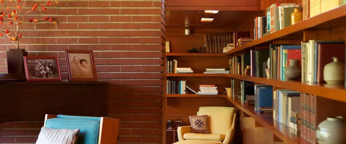 A cozy reading nook with a brick wall, a wooden bookshelf filled with books, a small armchair, and a framed photograph.