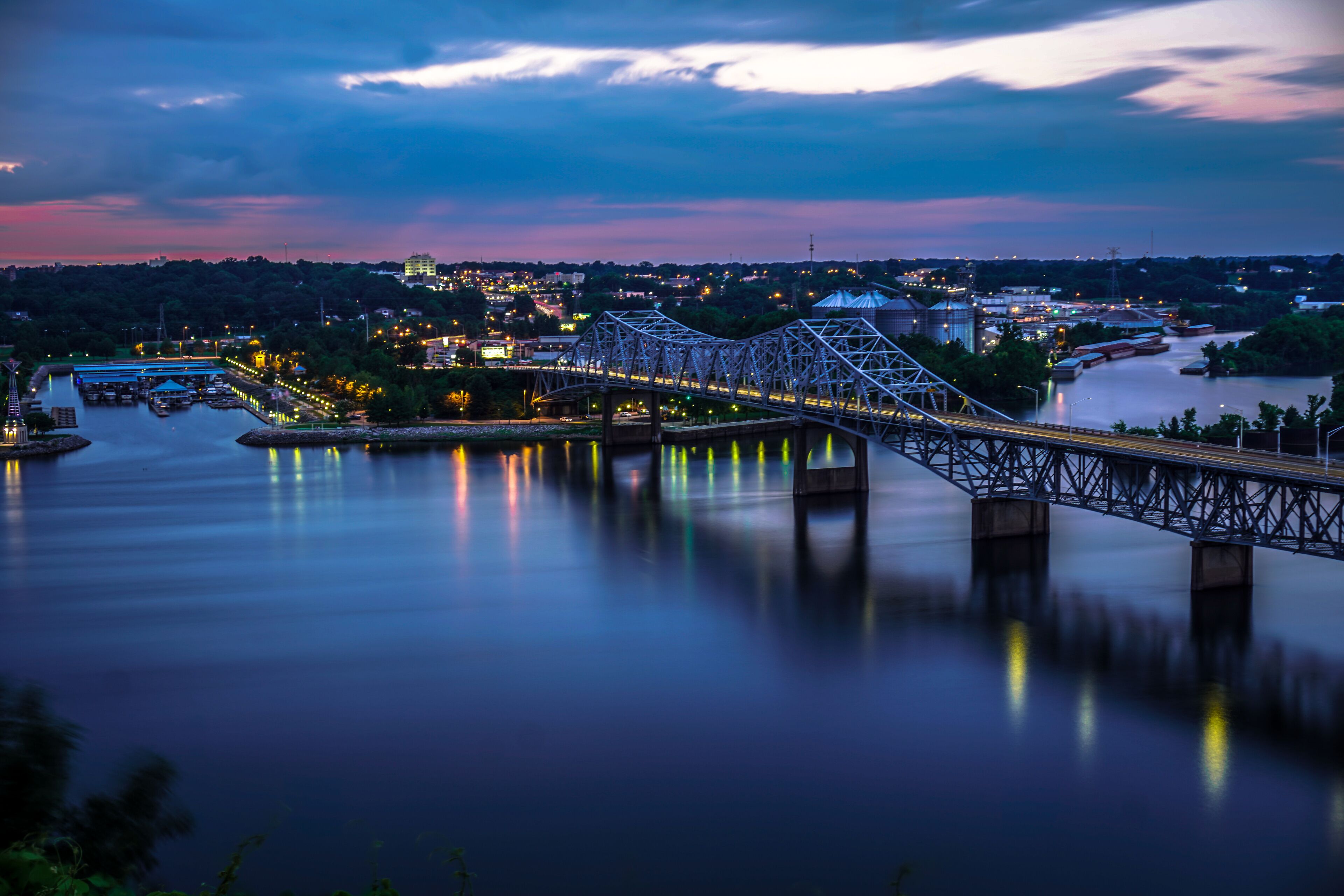 A steel bridge over a calm river at dusk with city lights in the background.