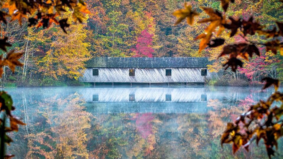 Covered bridge in the fall, As mist rises from the calm waters, fall colors are reflected around the covered bridge.