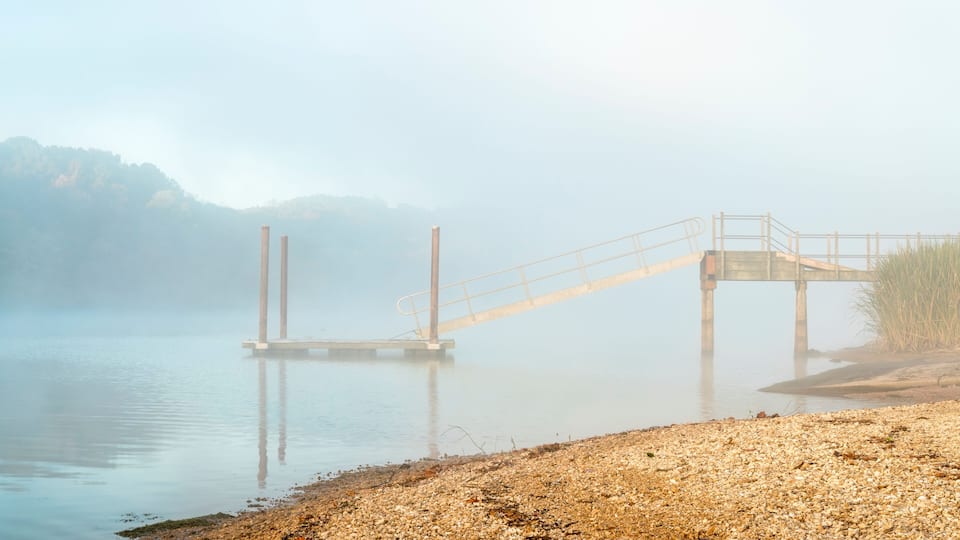 A foggy lake with a wooden pier extending into the water, surrounded by a pebbly shore and distant hills.
