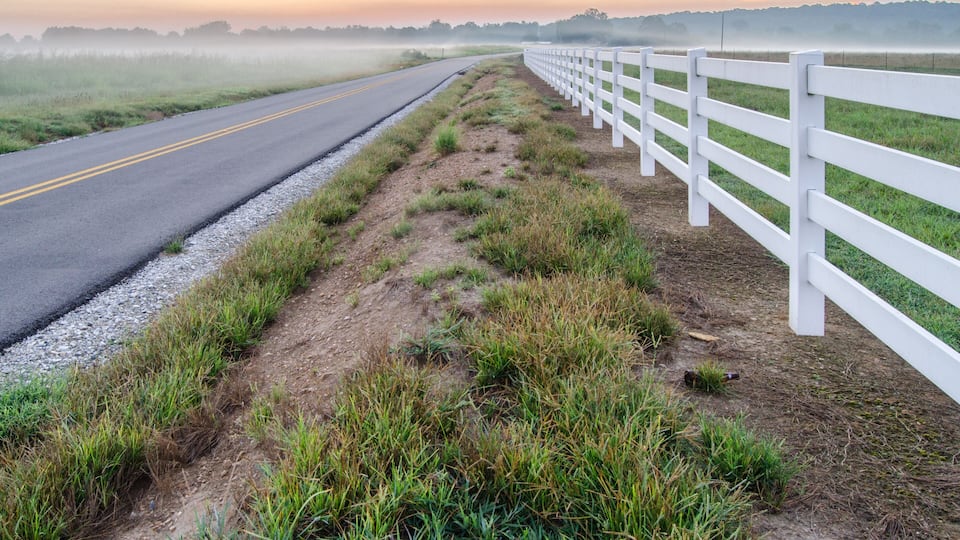 A rural road with a white fence on one side and a grassy field on the other.